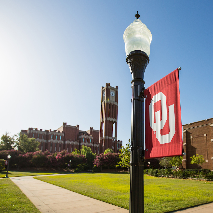 In the front of the image, a crimson flag with white OU letters hangs on a light pole with Bizzell Memorial Library and the Clock Tower in the background.